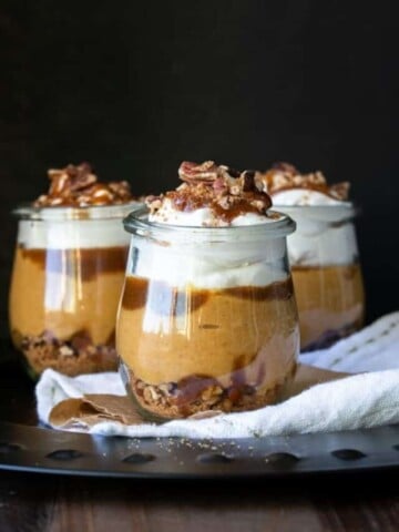 Three glass jars layered with cookies, pumpkin, caramel and whipped cream.