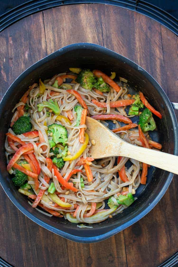 Wooden spoon mixing noodles and veggies in a pan