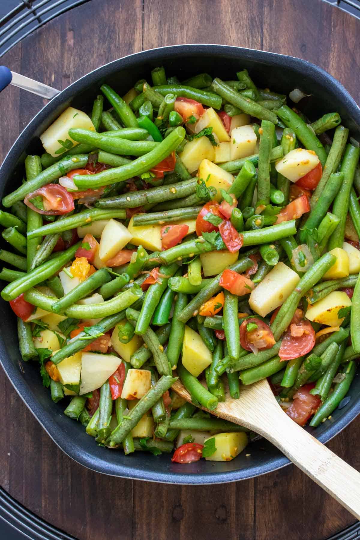 Wooden spoon stirring green beans, potatoes and tomatoes in a pan