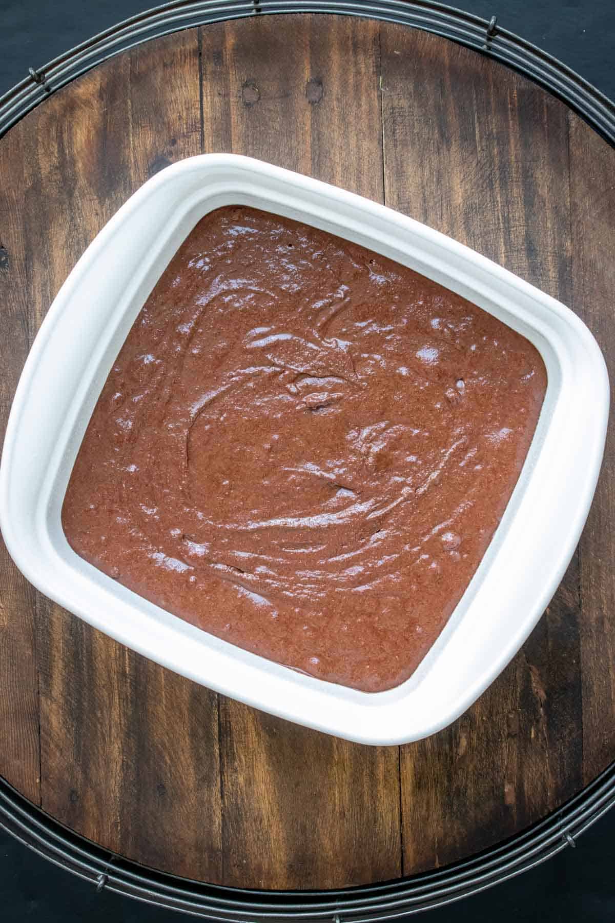 White baking dish on a wooden surface with raw brownie batter in it.