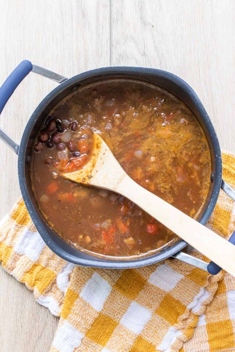 Wooden spoon mixing soup with black beans, peppers and onions in a black pot