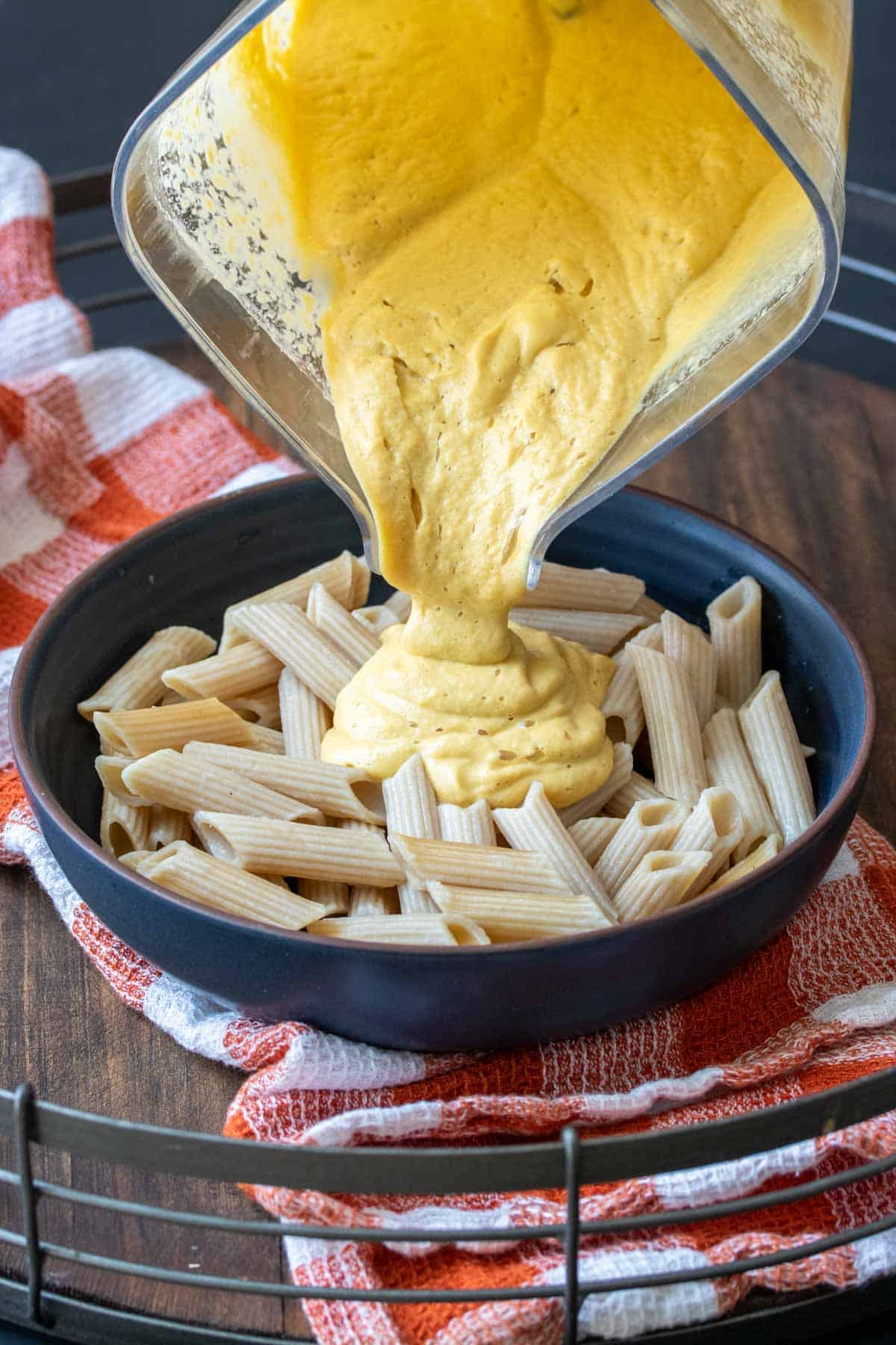 A blender pouring creamy butternut squash sauce over a bowl of penne pasta