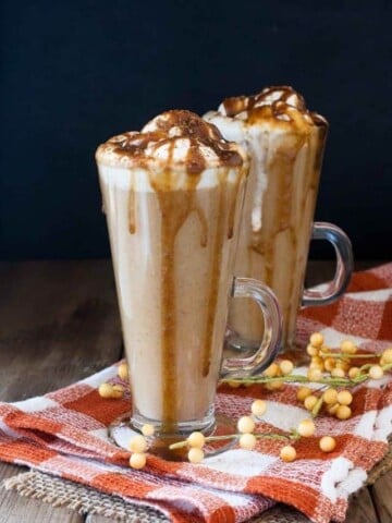 Two tall glass mugs with pumpkin lattes inside covered in whipped cream and caramel