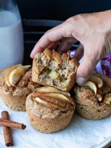 Hand grabbing a half eaten apple muffin off a pile of more muffins.