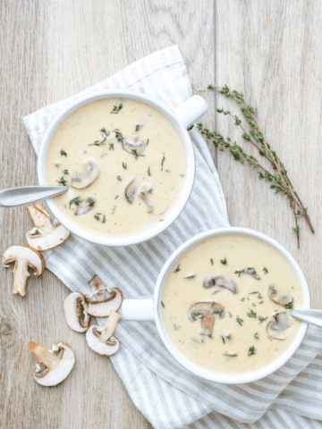 Cream of mushroom soup in two white bowls on a striped towel next to thyme and mushrooms.