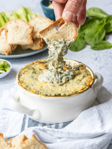 Bread piece dipping into a baked spinach artichoke dip.