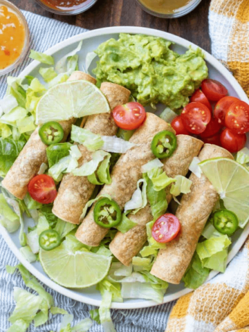 A grey plate with lettuce and taquitos sitting on top with guacamole and tomatoes.