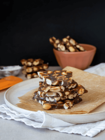 Pile of peanut brittle pieces on a piece of parchment folded up on a plate sitting on a white napkin.