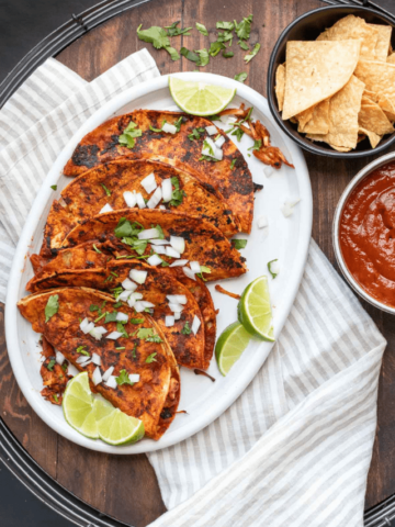 A white plate with five pan fried tacos covered in red sauce and topped with chopped onions.