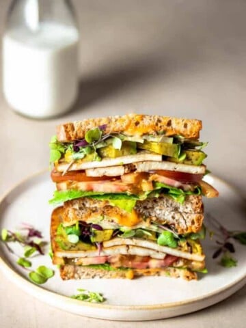 A tofu and veggie sandwich that has been cut in half and stacked on a tanish plate in front of a glass of milk.