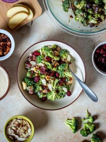 A creamy broccoli salad with cranberries in a tan bowl with a brown rim surrounded by ingredients and more salad.