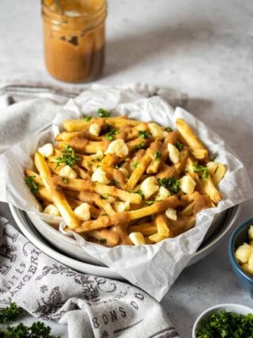 A stack of white bowls with parchment paper and poutine style french fries in them next to a towel and jar of gravy.