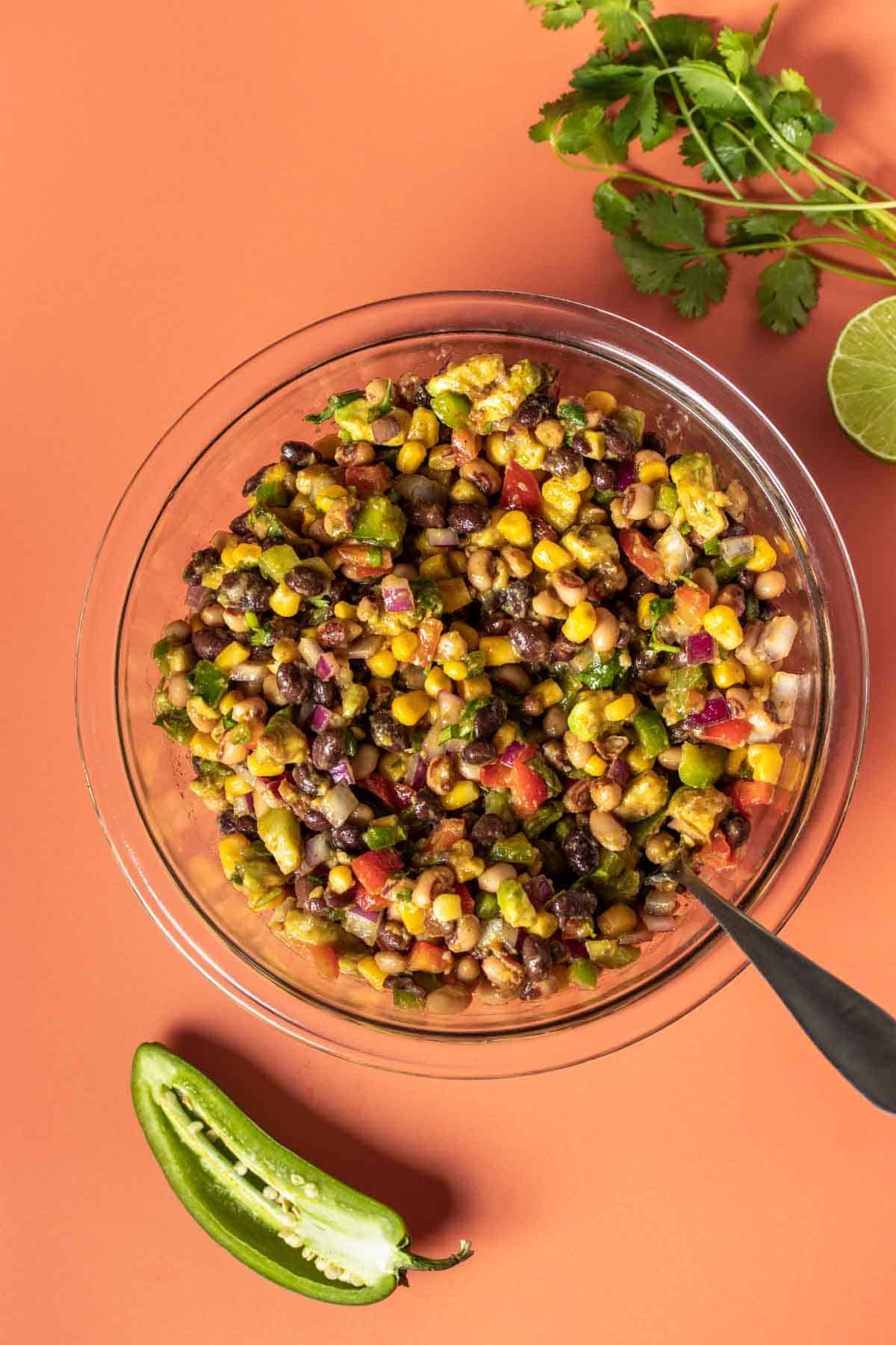 A glass bowl with a mixed cowboy caviar salad in it and a spoon on an orange background.