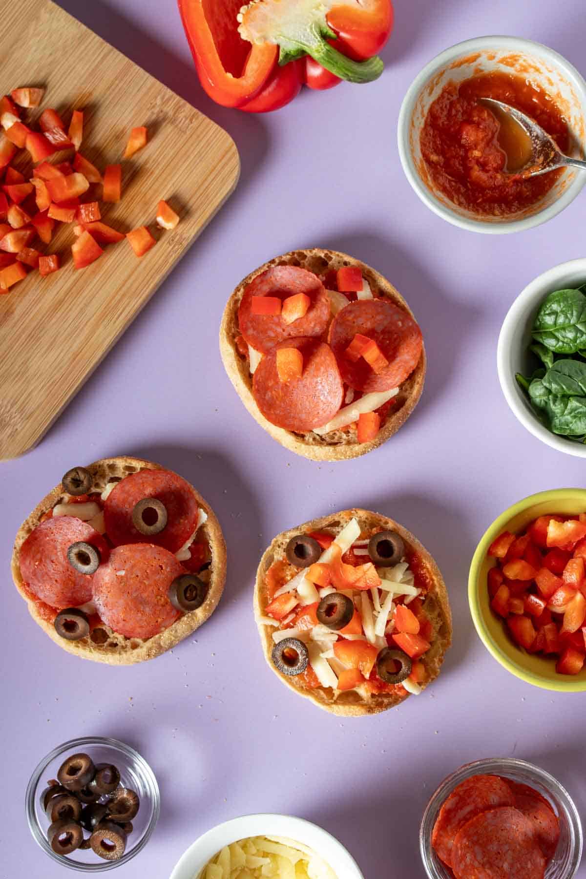 Cheese, pepperoni and olive English muffin pizzas on a purple surface with ingredients around them.