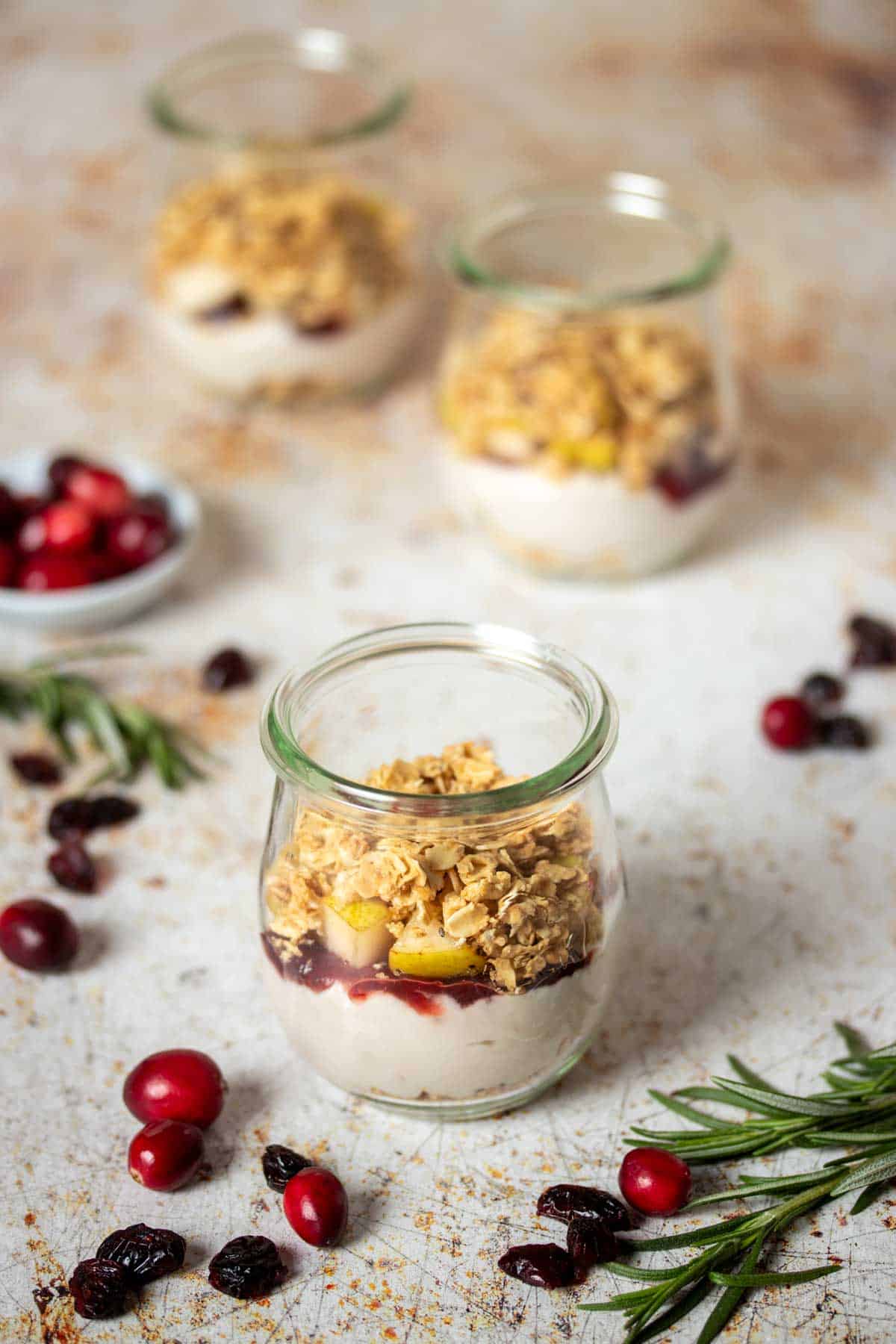 A speckled tan surface with three glass jars in front of each other layered with yogurt, jelly and granola.